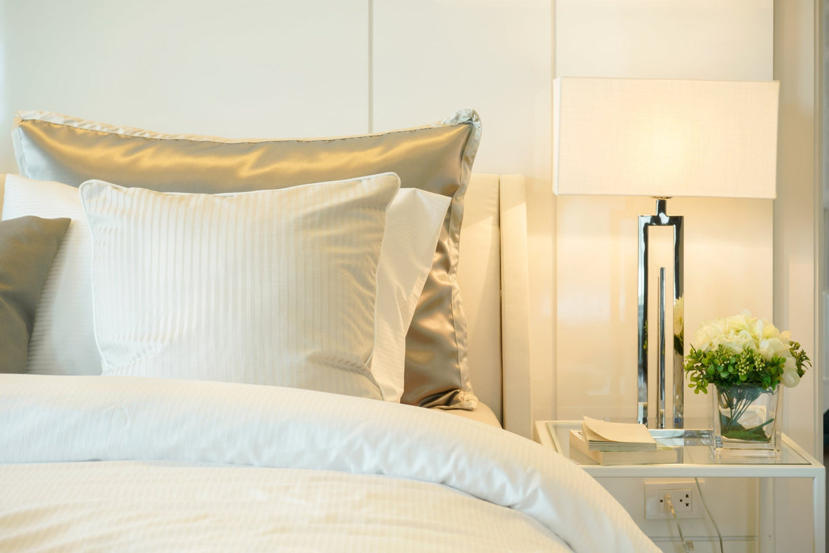 Warm bedroom with layered white bedding, a champagne-colored pillowcase, bedside lamp, and flowers