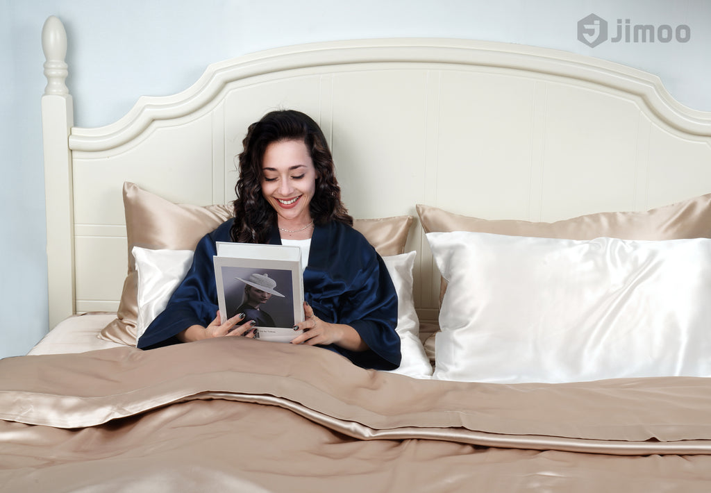 Woman smiling in bed with ivory white and champagne silk pillowcases, reading on a relaxed morning.