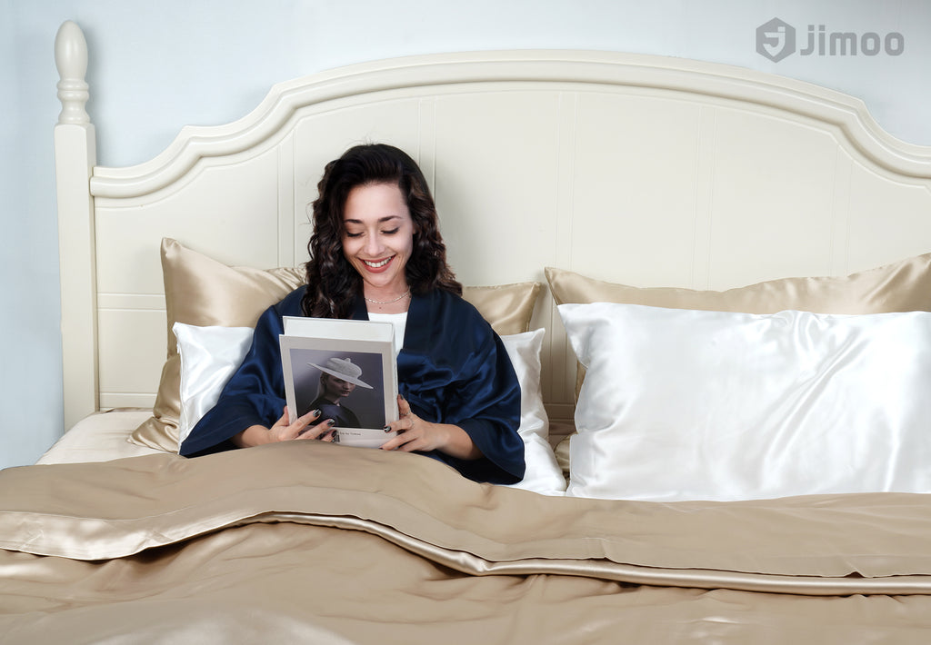 Woman smiling in bed with beige silk pillowcases, enjoying a relaxed morning read.