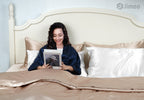 Woman reading in bed with pure white and champagne silk pillowcases, smiling on a relaxed morning.